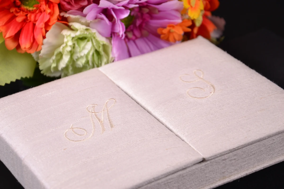 Pale invitation book folio with embroidered initials beside colourful wedding flowers
