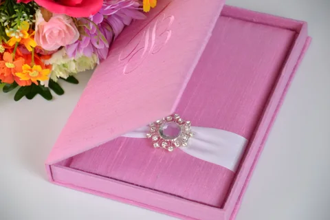 Open pink invitation box with inner card panel, ivory ribbon and a jeweled accent