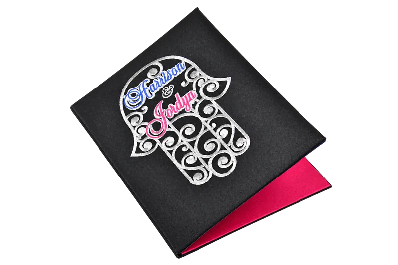 Black invitation folio with silver ornamental embroidery, blue and pink names, and a pink interior