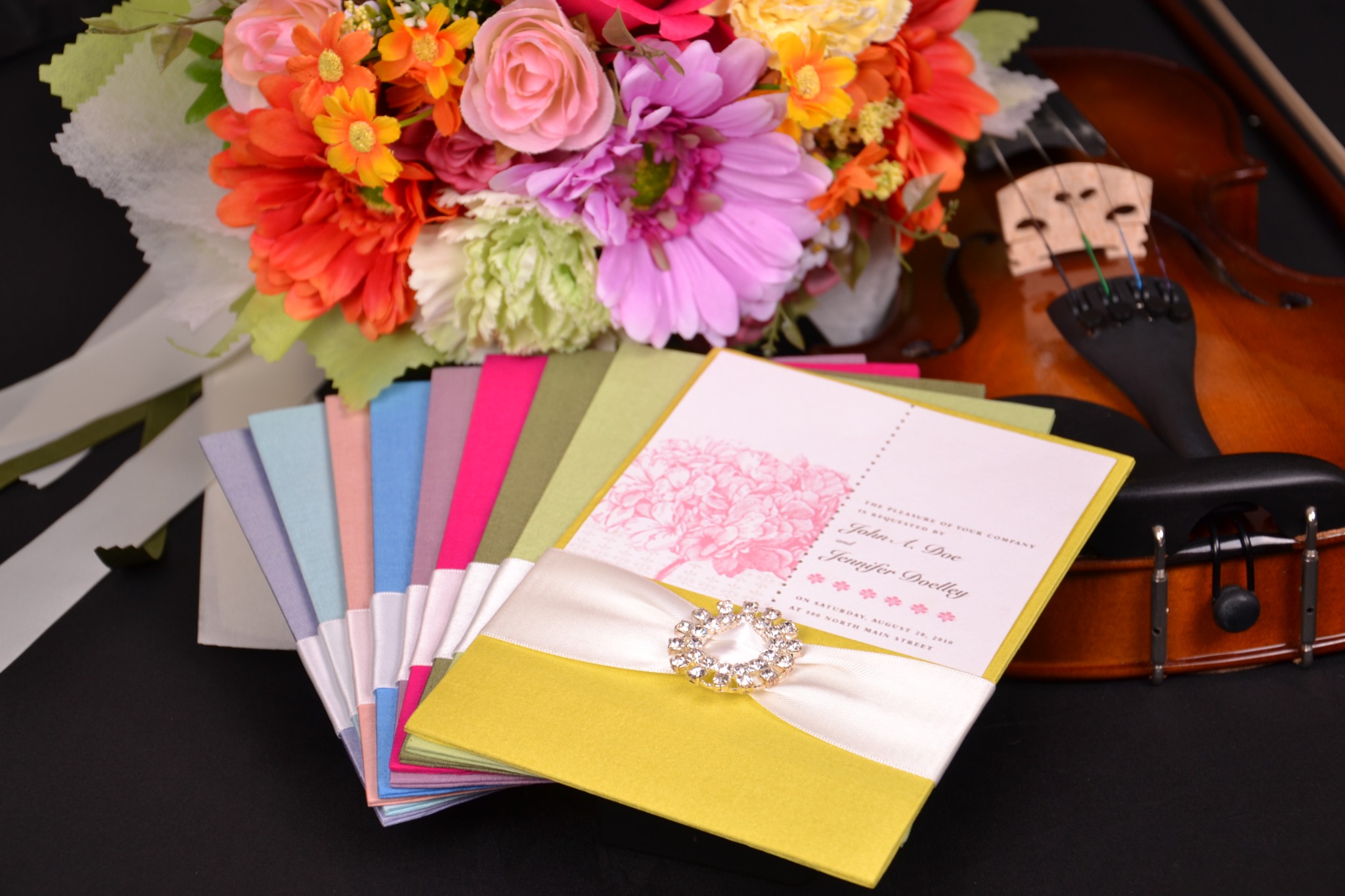 Custom silk wedding invitation folios in multiple colors with satin ribbons and jeweled embellishment beside wedding flowers and a violin